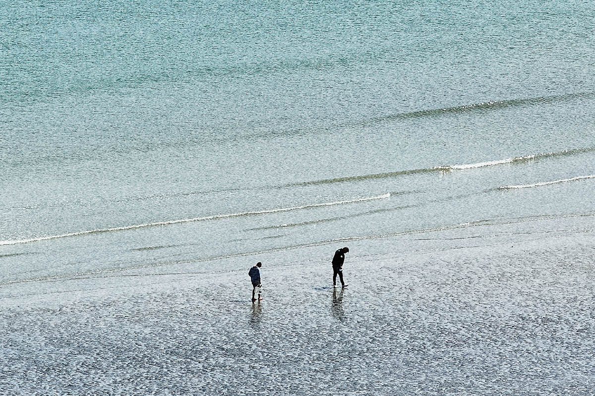 The blurred silhouettes of two men searching for seashells at the water's edge on a beach in Brittany in the summer, from the series Another summer by Emmanuel Pineau