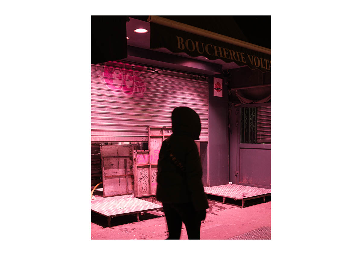 A young woman walks at night in the pink light of a closed shop, from the series I will be around by Emmanuel Pineau