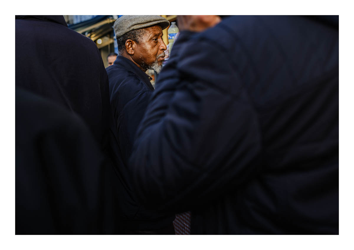 A man waits his turn in front of a shop on Paris Street, from the series I will be around by Emmanuel Pineau