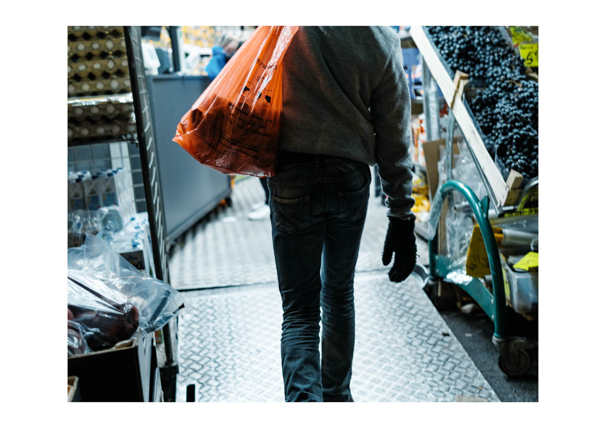 A man carrying an orange bag enters a shop on Rue de Paris one winter evening., from the series I will be around by Emmanuel Pineau