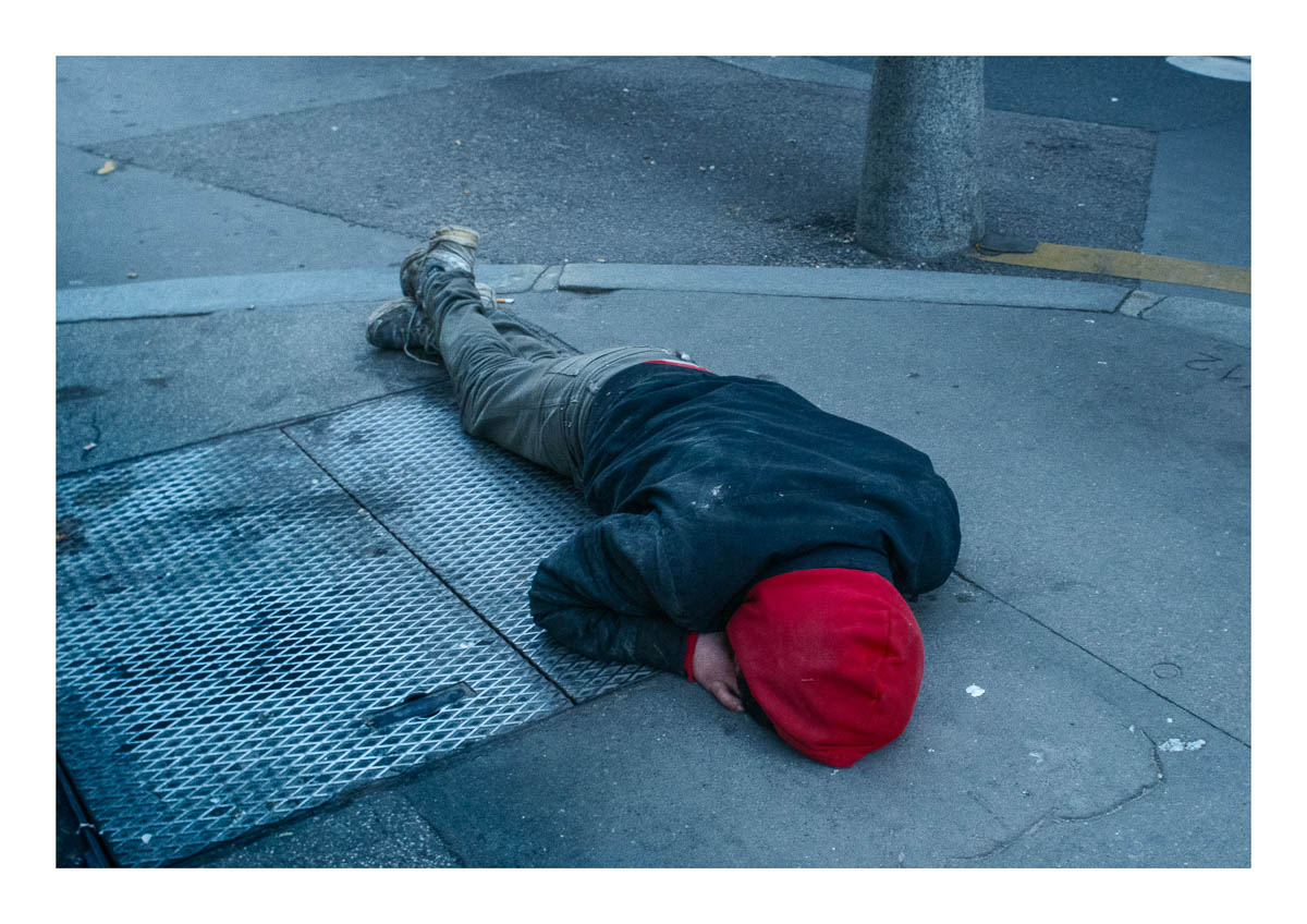 A homeless man is lying on the ground, his face hidden by his red hood., from the series I will be around by Emmanuel Pineau