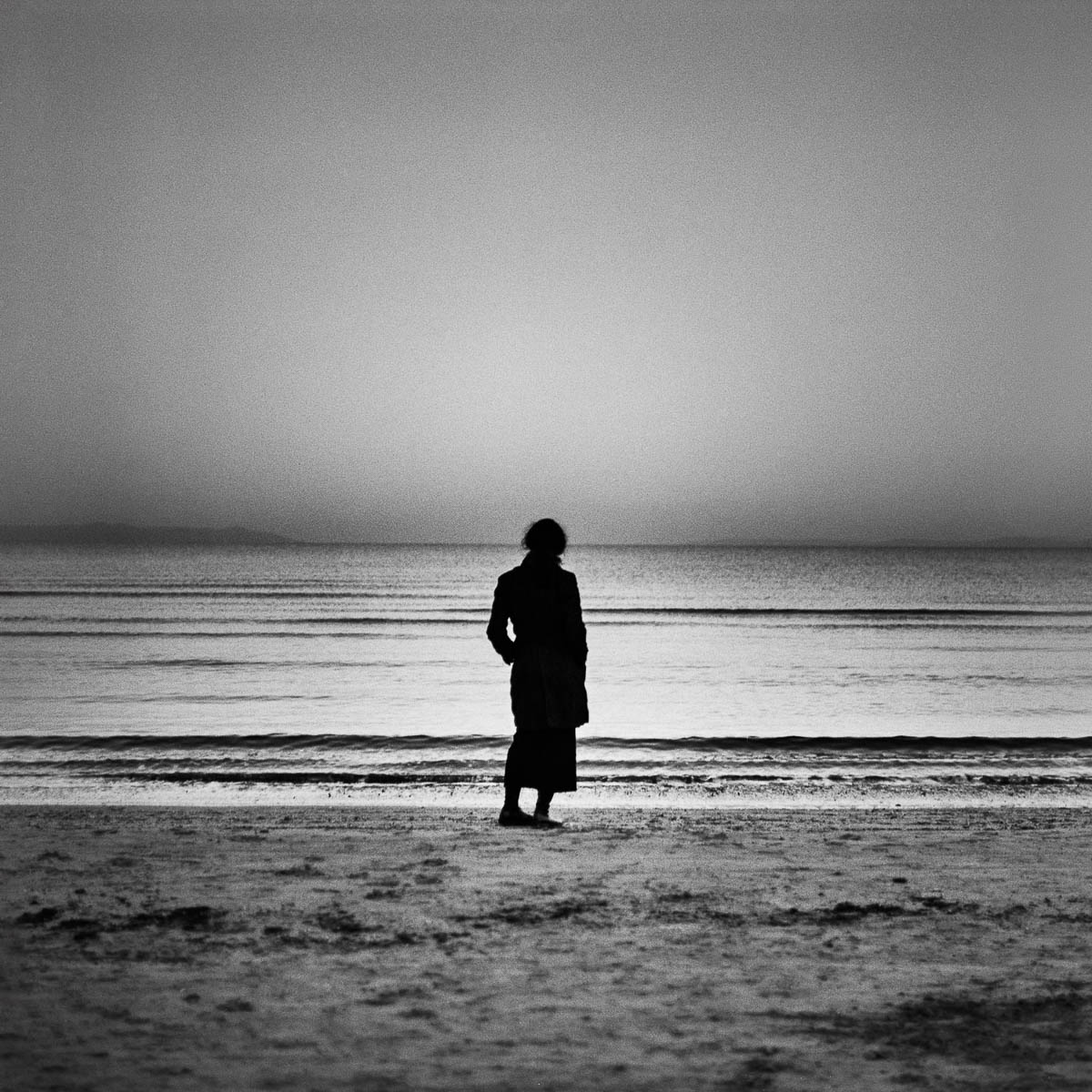 The silhouette of a woman seen from behind, wearing a dark coat, looking out at the sea, from the series Les limites du territoire by Emmanuel Pineau