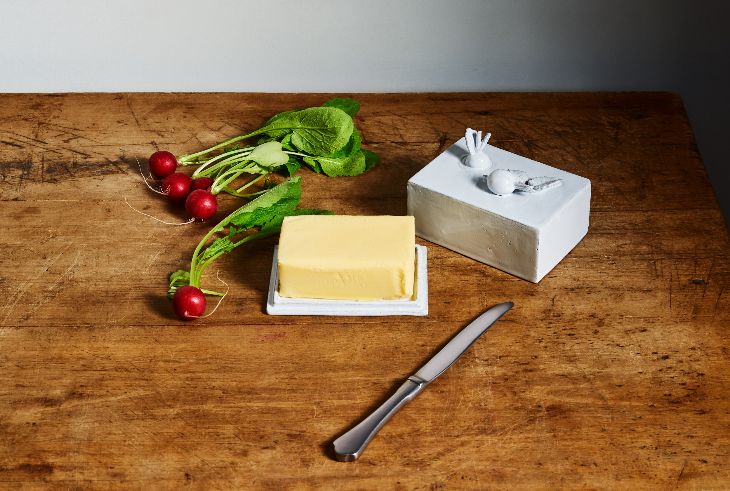 White ceramic butter dish by Astier de Villatte on a rustic wooden table with a bunch of radishes, still life photography by Emmanuel Pineau