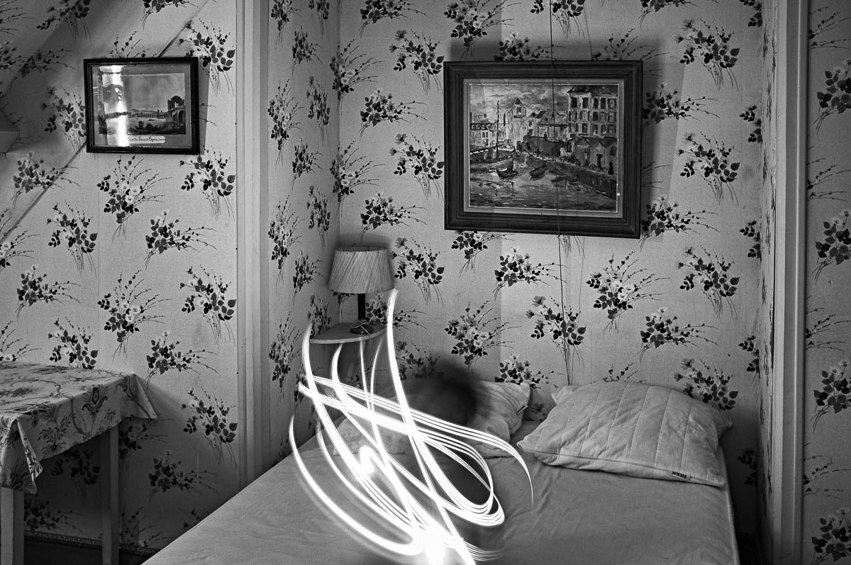 A child sitting on a bed in a vintage bedroom is having fun doing light painting, from the series Out of disorders by Emmanuel Pineau