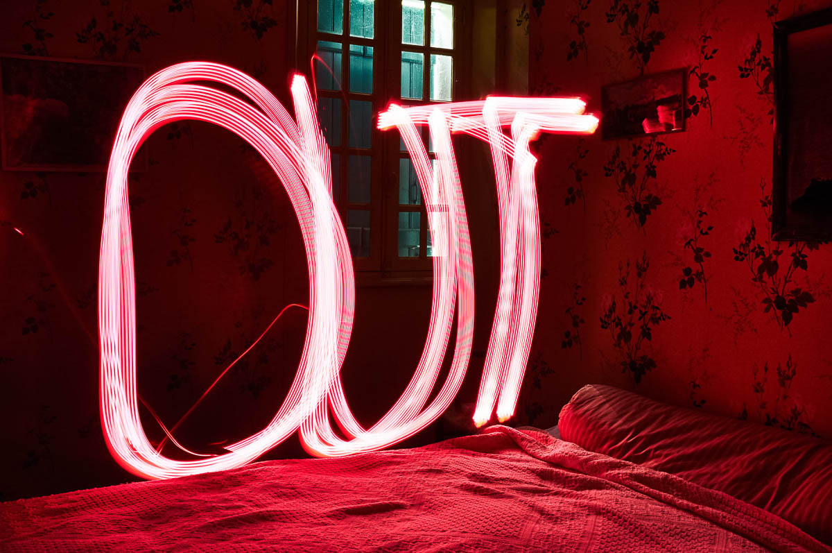 Vintage bedroom with the word OUT written in red light painting above the bed, from the series Out of disorders by Emmanuel Pineau