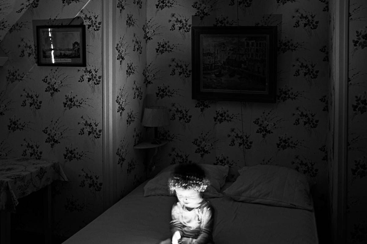 A little boy sitting on the bed in a vintage bedroom plays with a flashlight, from the series Out of disorders by Emmanuel Pineau