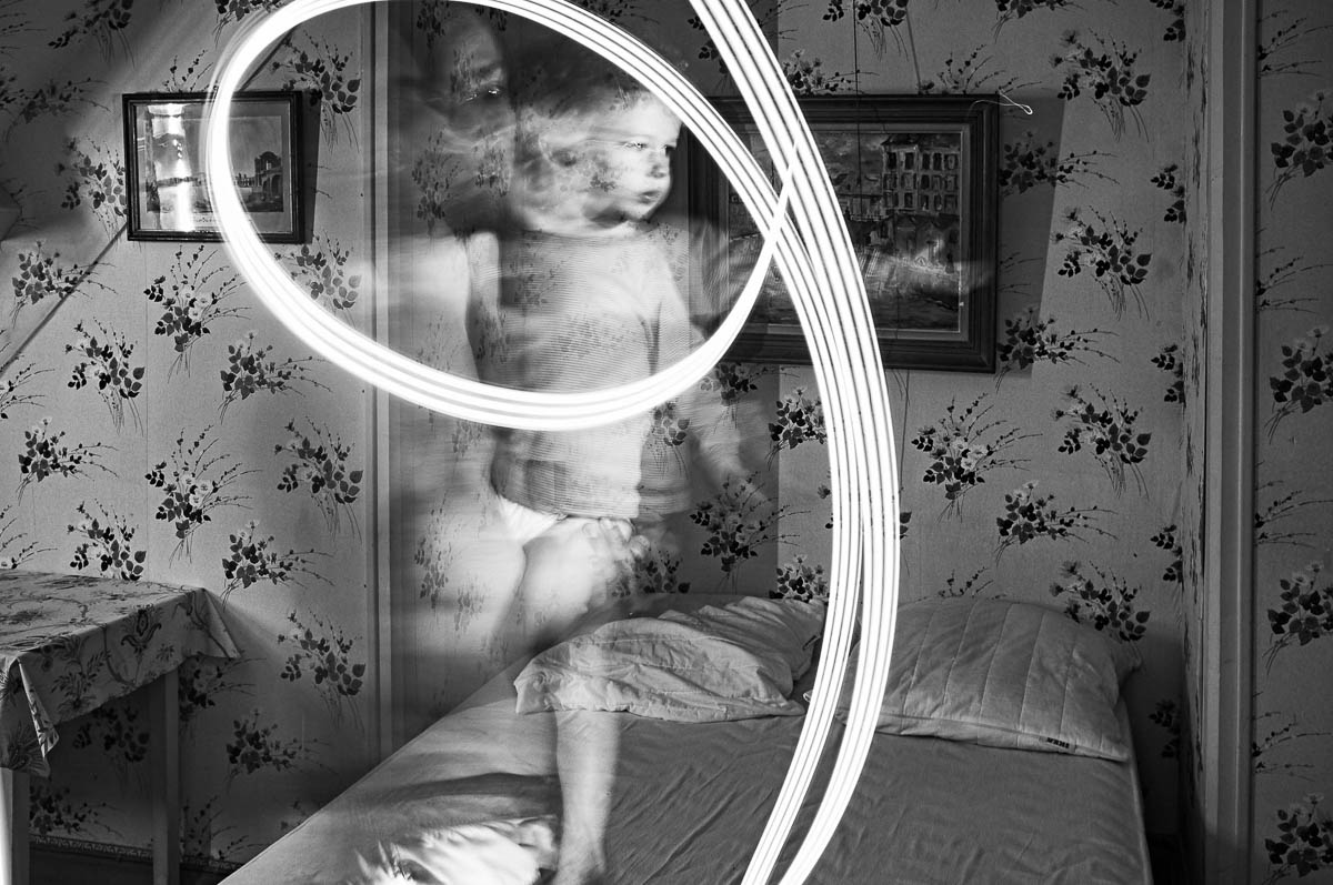 A father carrying his son in his arms plays with a flashlight in a vintage bedroom, from the series Out of disorders by Emmanuel Pineau