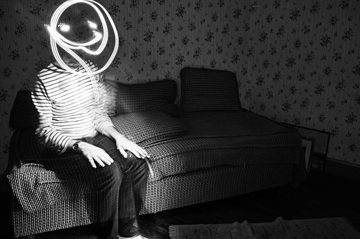 Self-portrait of Emmanuel Pineau sitting on a vintage sofa with a smiley face drawn in light painting on his face, from the series Out of disorders by Emmanuel Pineau