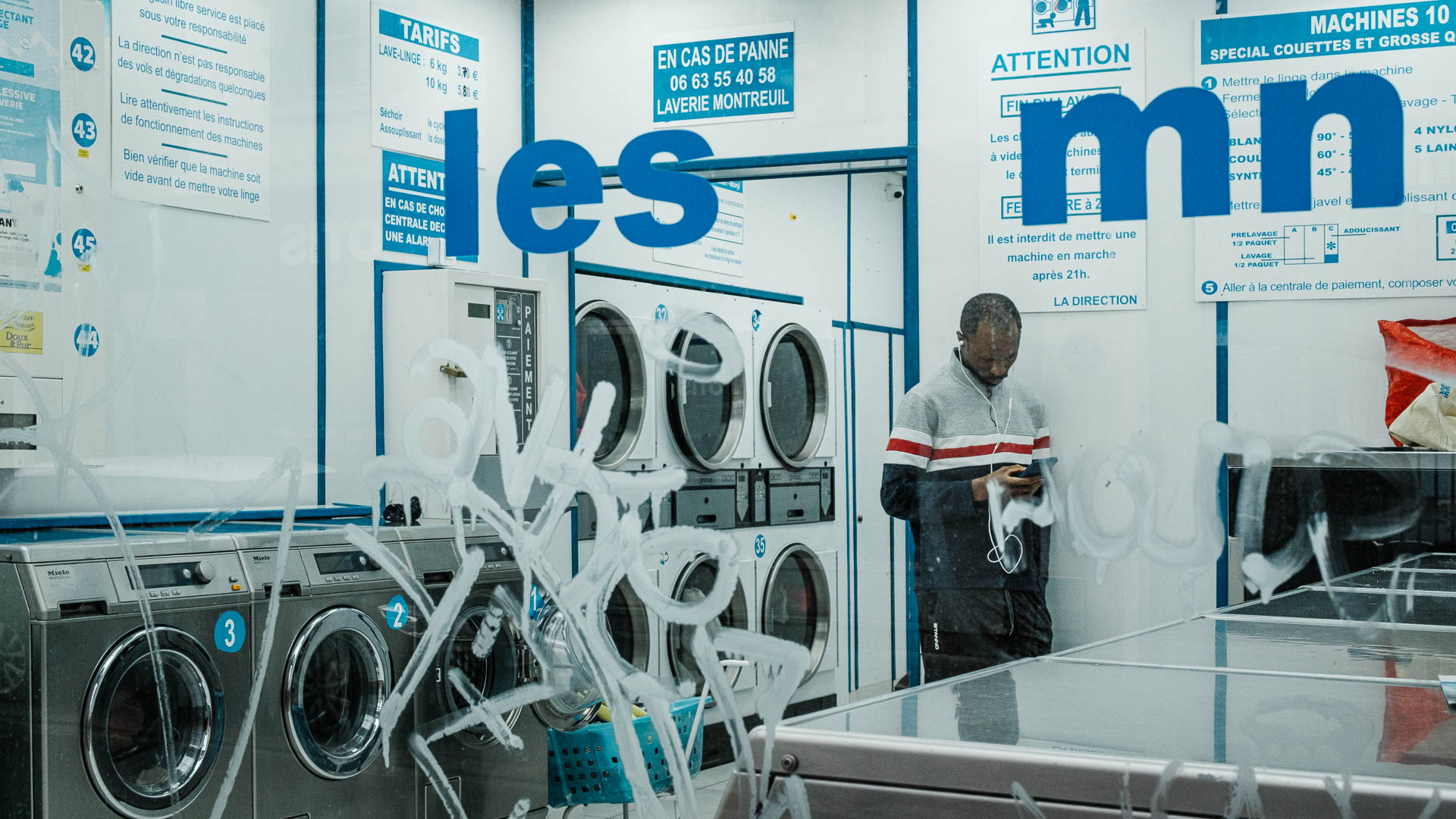 A man alone in a laundromat at night, looking at his phone, from the series I will be around by Emmanuel Pineau